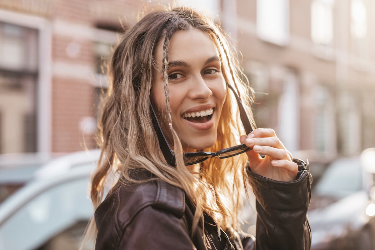 Woman standing in the street holding her sunglasses by her mouth and smiling